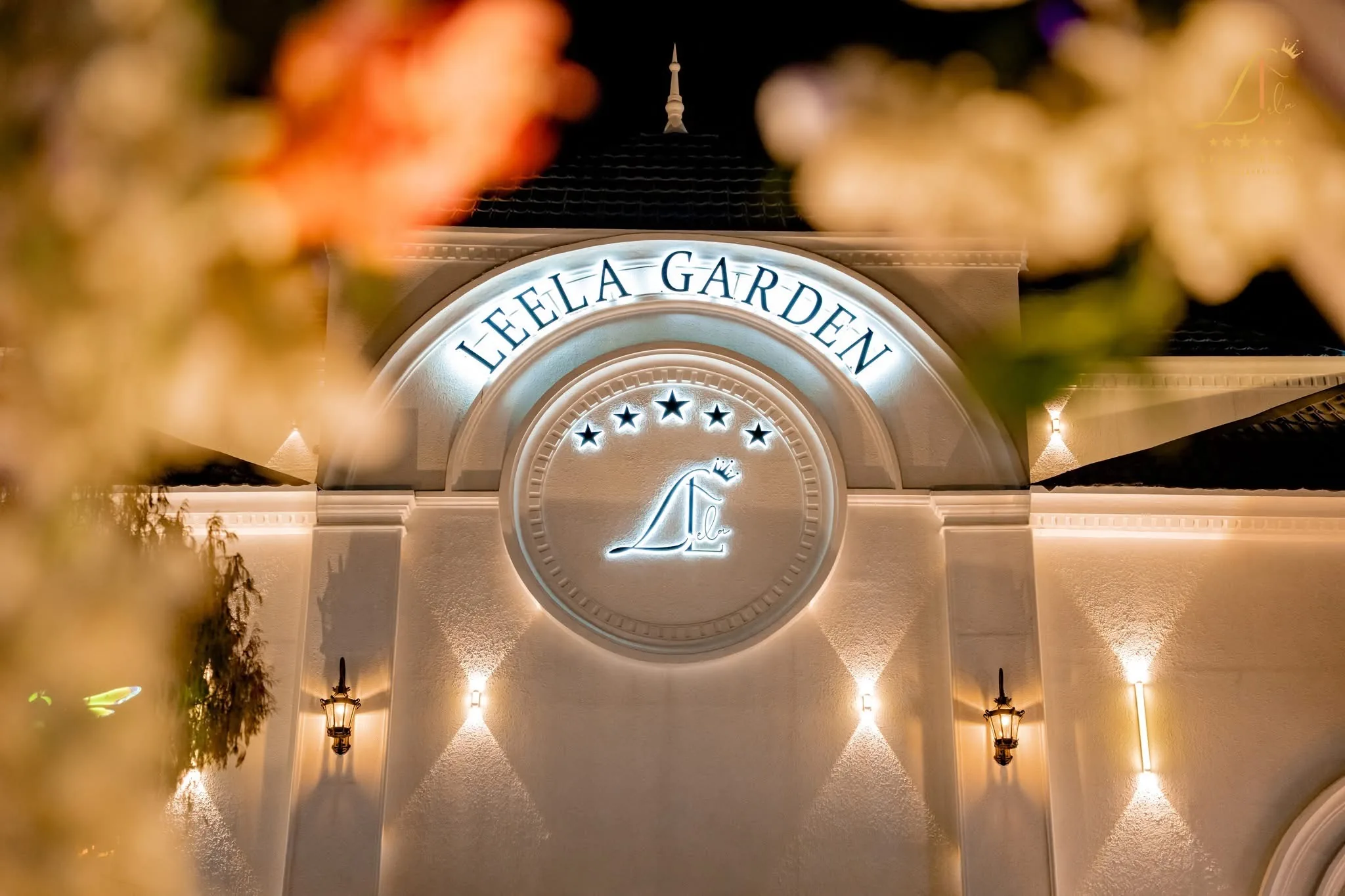 The illuminated Leela Garden crest above the entrance at night, framed by softly blurred flowers.